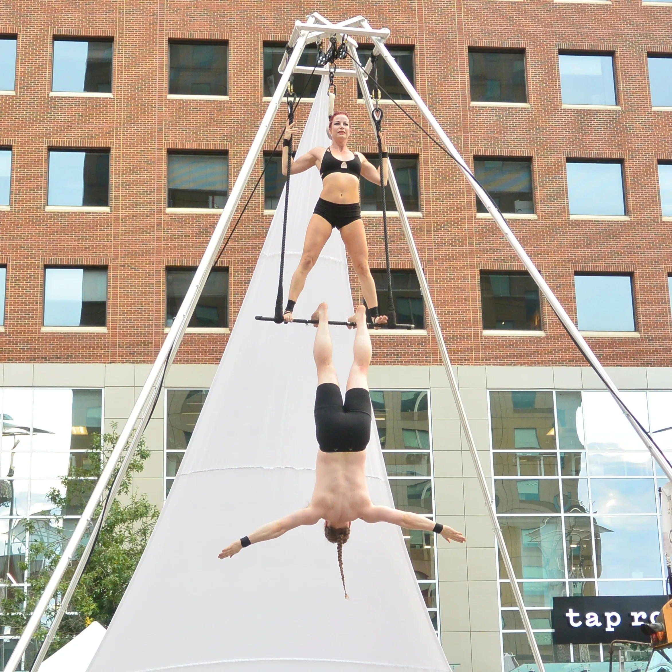 An aerialist hangs inverted beneath her partner on an outdoor rig